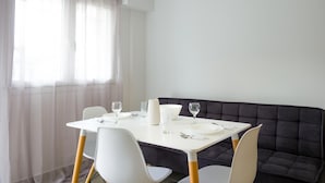 A dining area with a white table set for two, a dark cushioned bench, and sheer curtains. - Elounda Olithos Apartments 2 (Agios Nikolaos)