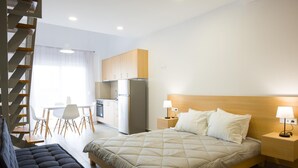 A modern hotel room with a large bed, a small sofa, a wooden headboard, a nightstand with a lamp, a kitchenette, and a staircase. - Elounda Olithos Apartments 4 (Agios Nikolaos)