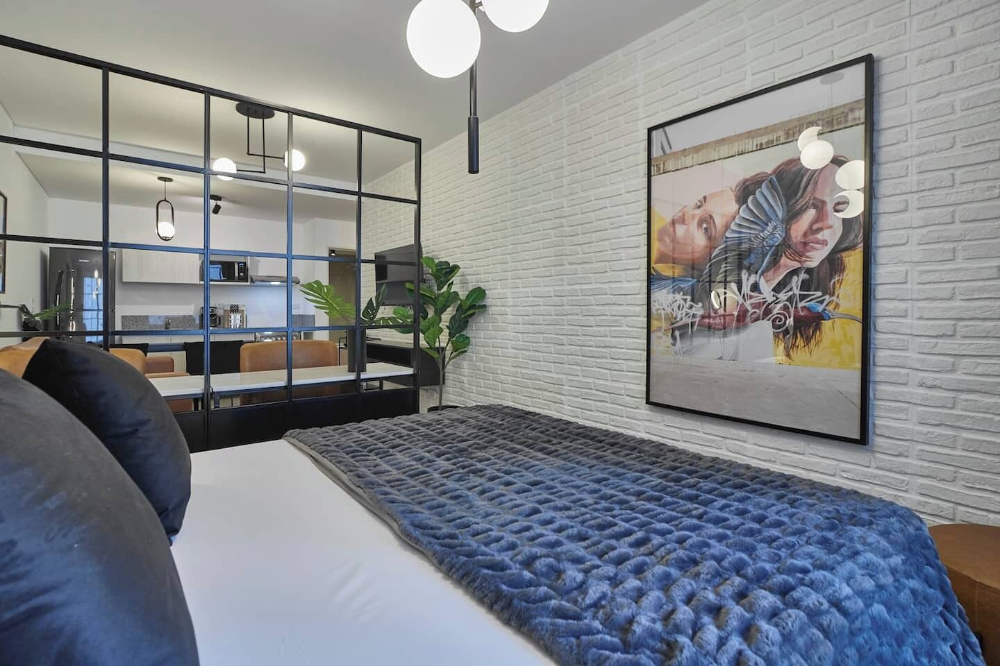 A modern bedroom with a large bed, a blue textured rug, a white brick wall, and a framed artwork.