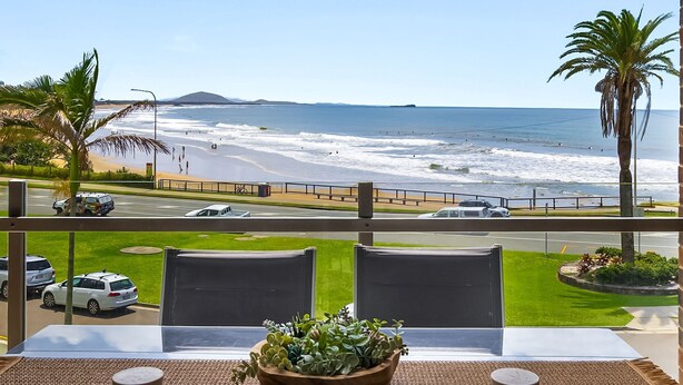 A balcony with a view of the beach and ocean. There are two chairs and a potted plant on the balcony.