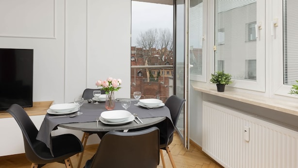 A dining area with a round table set for four, a flat-screen TV on the left, and a window with blinds overlooking a cityscape.