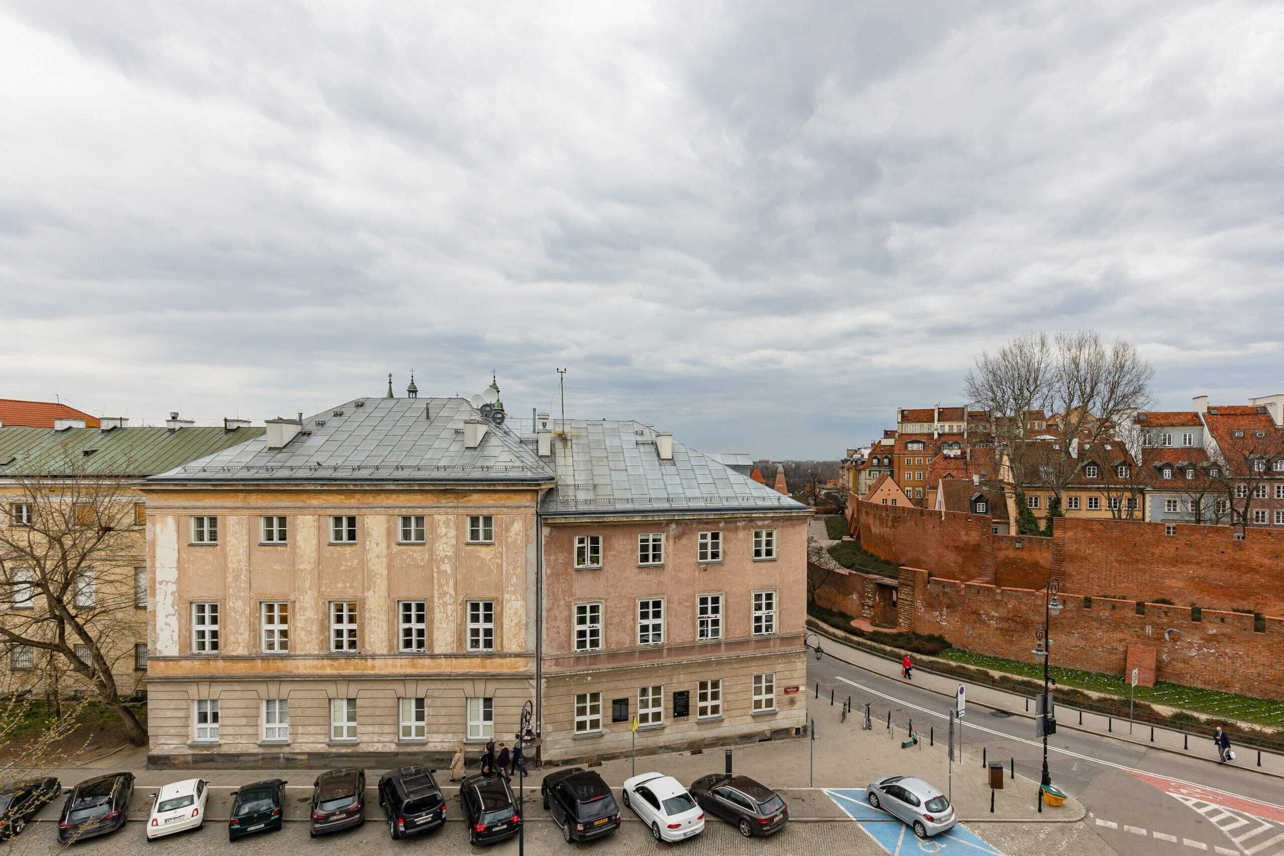 En historisk byggnad med ett markant torn, som ligger intill en mur av tegelsten och en asfalterad gata med parkerade bilar.