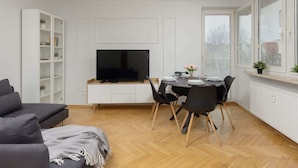 A modern living room with a sofa, dining table, and chairs. There is a TV mounted on the wall, a bookshelf, and a window with blinds.