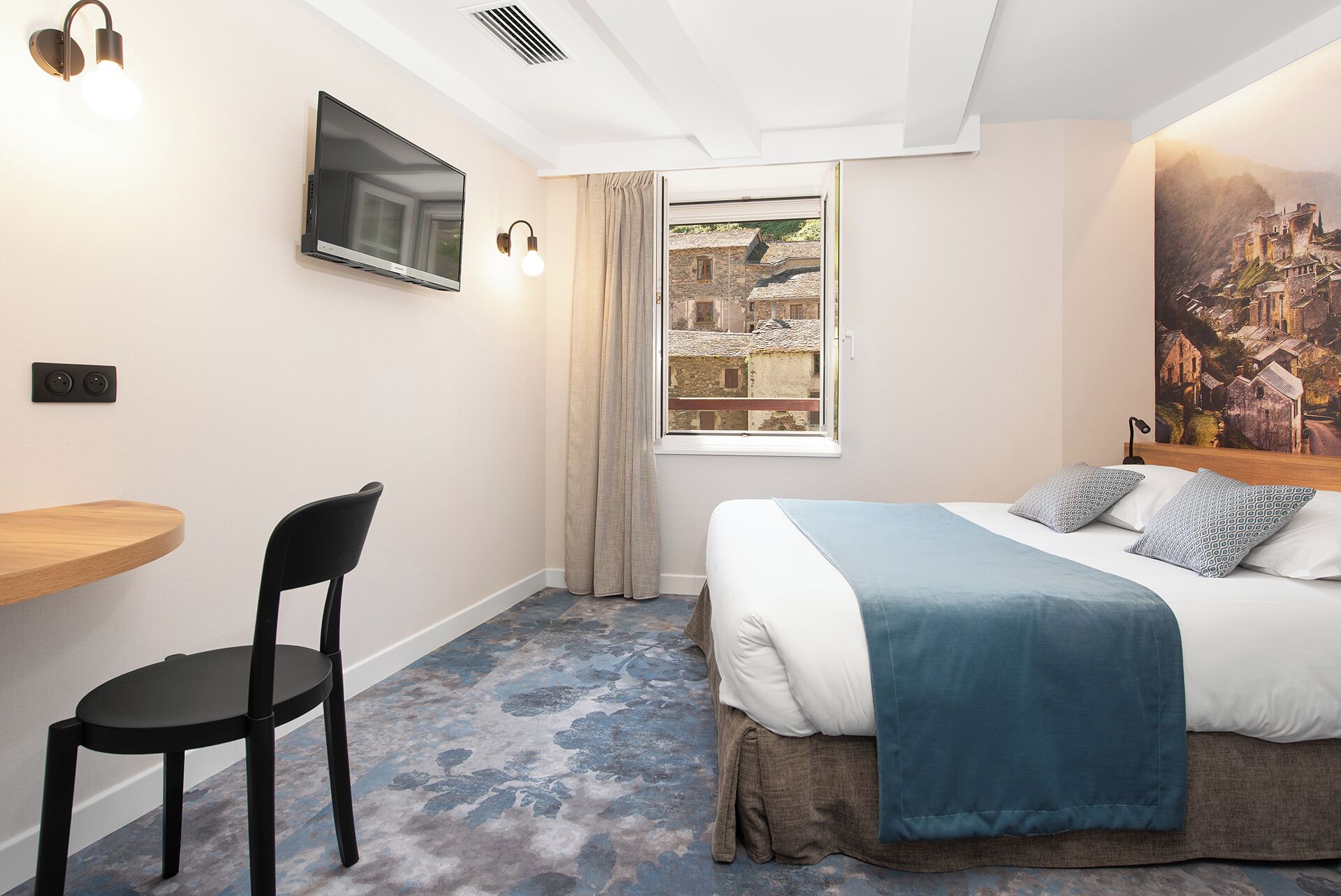 Een slaapkamer met een bed, een bureau, een stoel, een televisie en een raam met uitzicht op gebouwen.