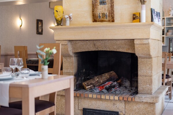 A stone fireplace with a fire, a dining table with chairs, and a wall-mounted light fixture. - Le Relays du Chasteau (Brousse-le-Chateau)