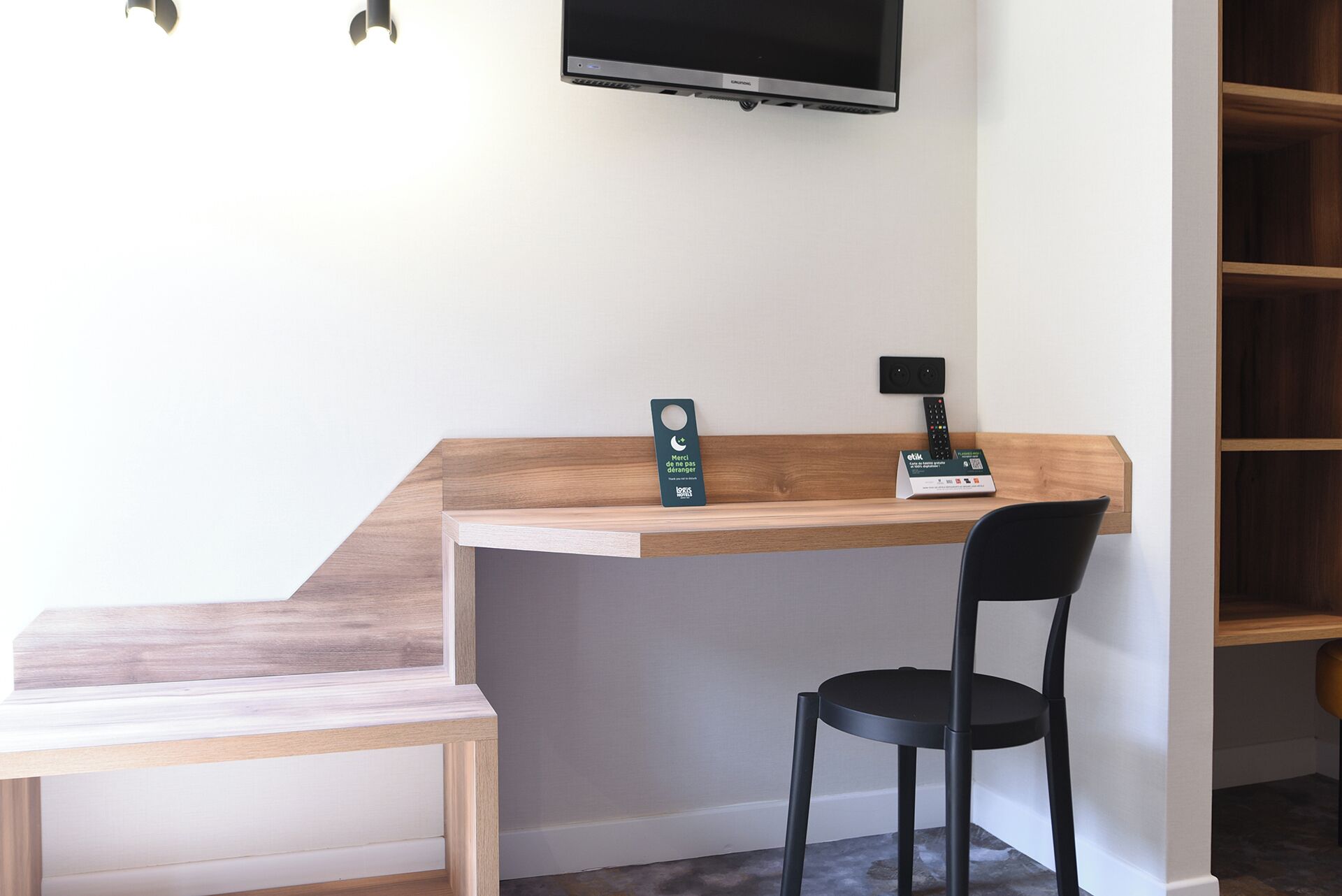 A hotel room with a wooden desk, a black chair, a TV mounted on the wall, and a remote control on the desk.