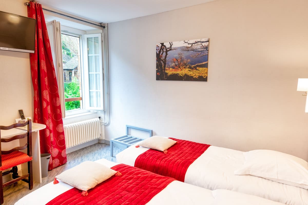 Et hotelværelse med to senge, et vindue med røde gardiner, en radiator, et lille bord og et billede på væggen.