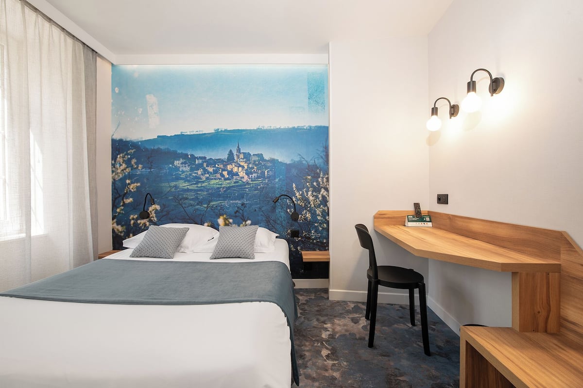 Et hotelværelse med en seng, et skrivebord, en stol og et vægmaleri af en by.