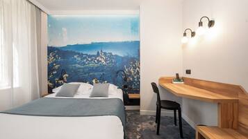 A hotel room with a bed, a desk, a chair, and a wall mural of a town.