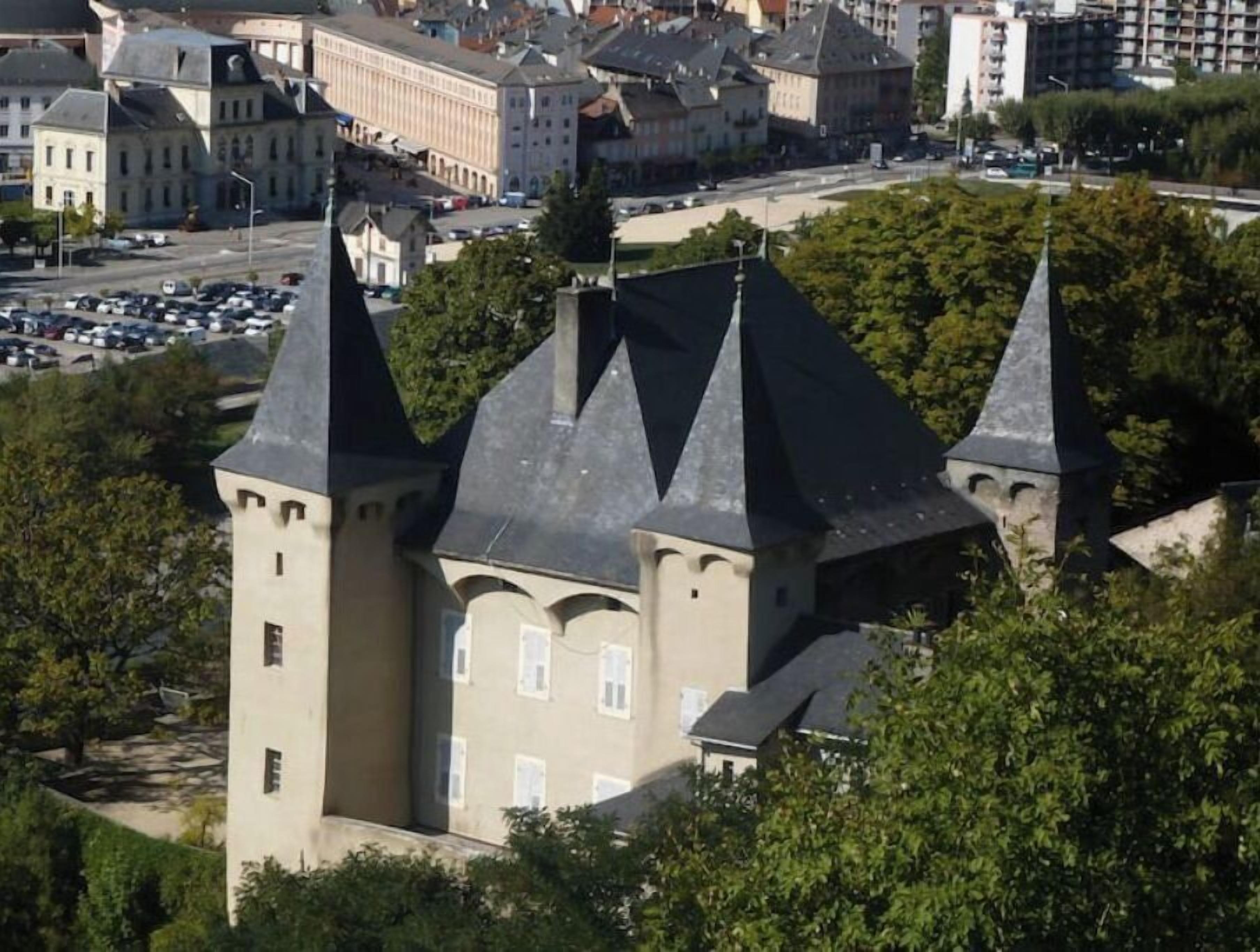 Ein schlossartiges Gebäude mit mehreren Türmen und einem zentralen Innenhof, umgeben von weiteren Gebäuden und einem Parkplatz.