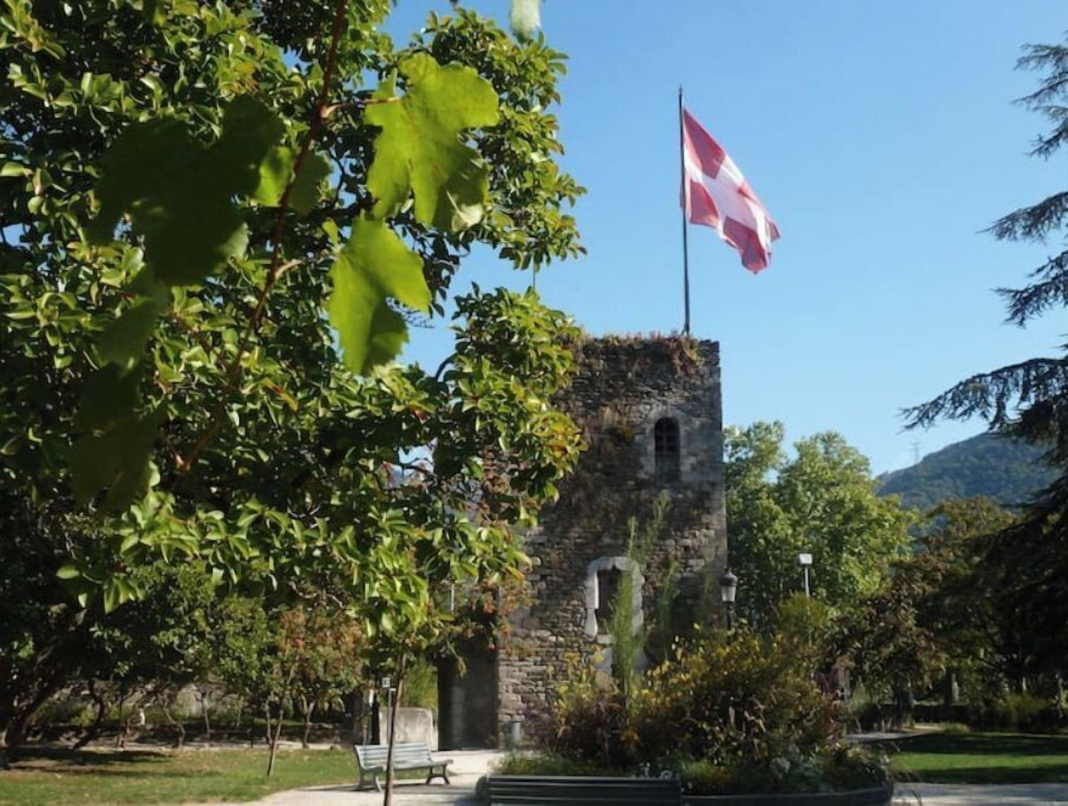 A stone tower with a Swiss flag, surrounded by trees and a clear sky.