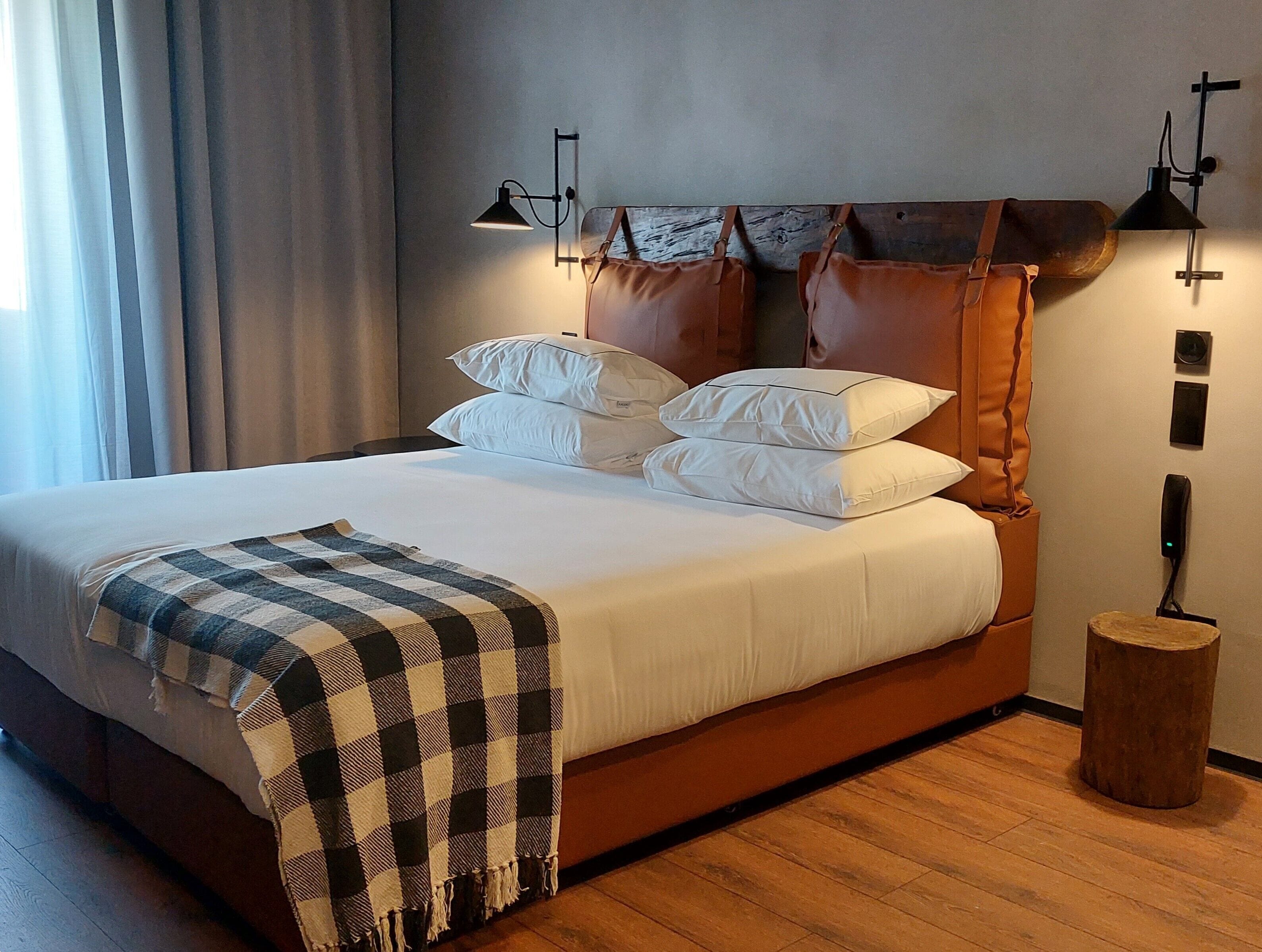 A neatly made bed with white linens and a checkered throw blanket. There are two wall-mounted lamps on either side of the bed.