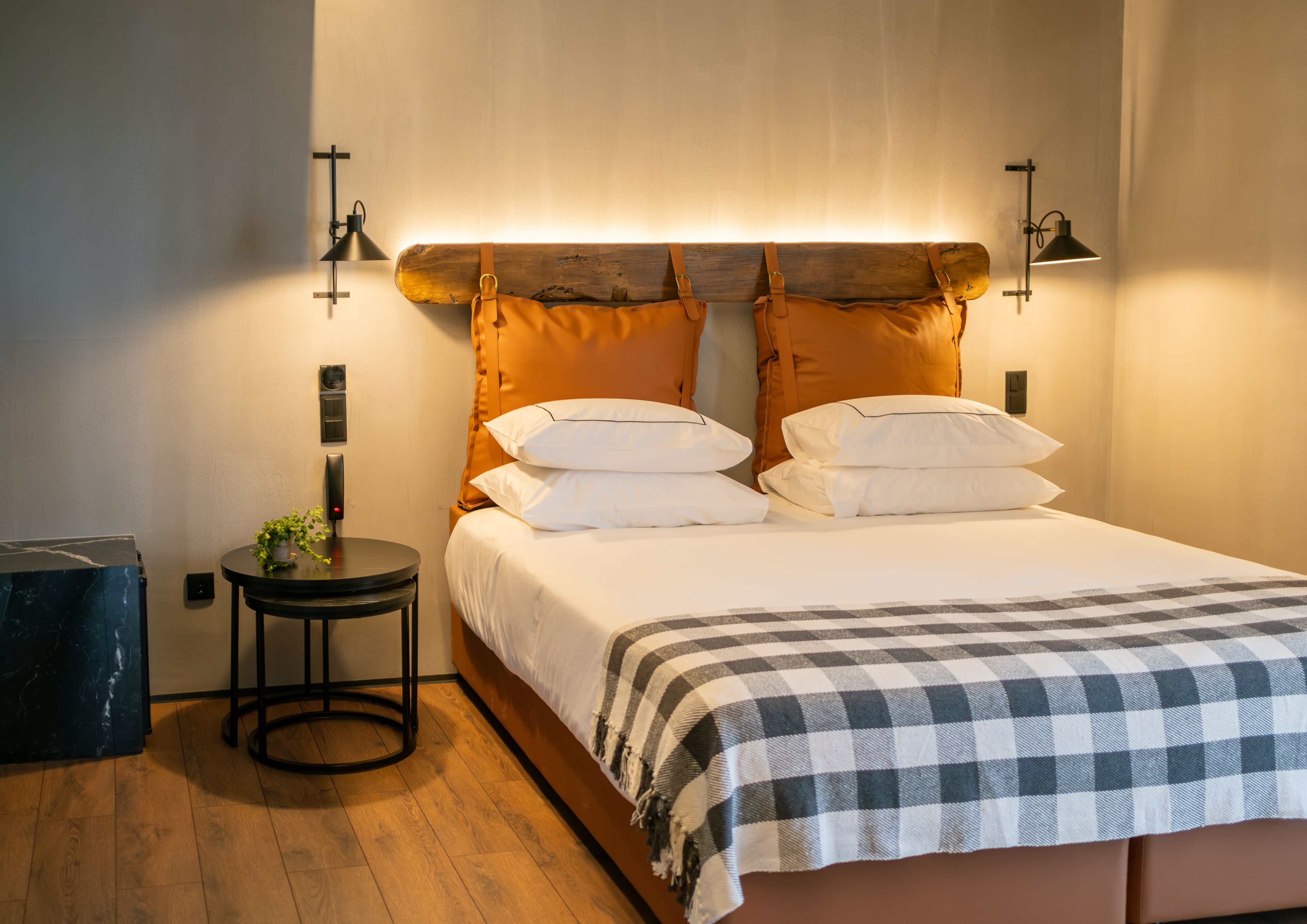 Un lit bien fait, recouvert d’une couverture à carreaux, une petite table ronde avec une plante, et deux lampes fixées au mur.