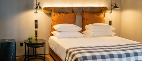 Una cama bien tendida con una manta a cuadros, una mesita redonda con una planta y dos lámparas de pared.