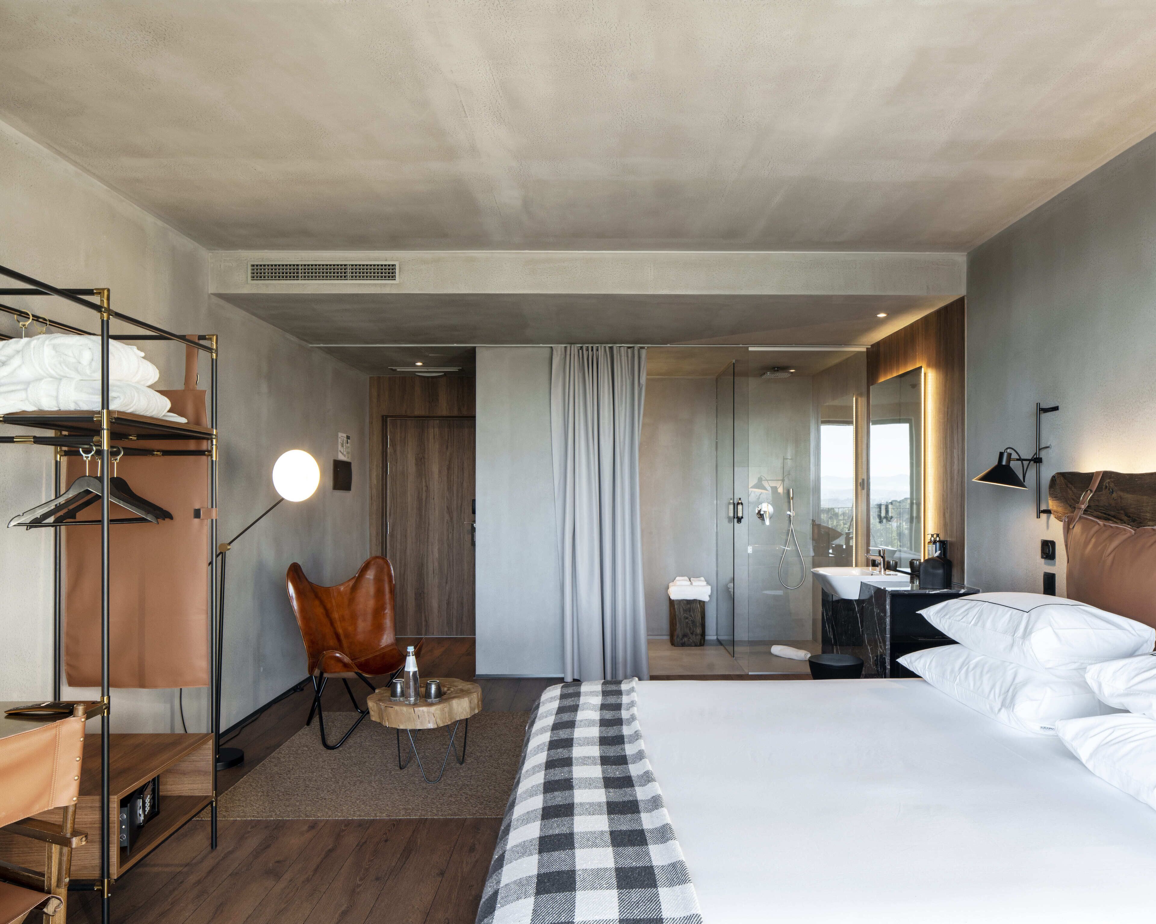 A modern hotel room with a large bed, a wooden side table, a leather chair, a desk, and a bathroom with a mirror and sink.