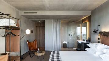 A modern hotel room with a large bed, a wooden side table, a leather chair, a desk, and a bathroom with a mirror and sink.
