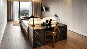 A modern hotel room with a large bed, a desk with a chair, a TV, and a view of the city through large windows. - Puro Dão Hotel & Spa (Nelas)