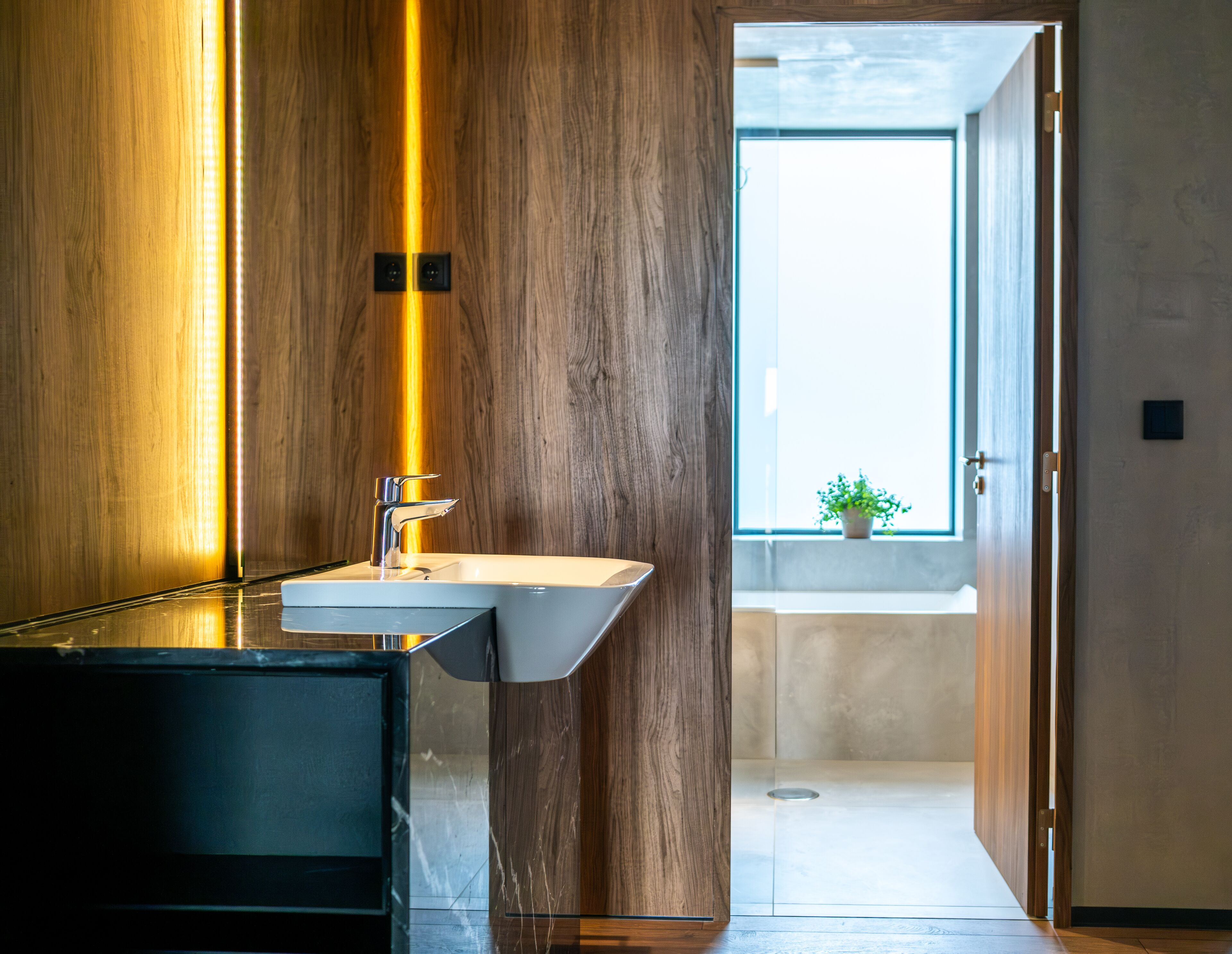 Ein modernes Badezimmer mit einer Holzwand, einem Waschbecken mit Chromarmatur und Blick auf einen Balkon mit einem Blumentopf.
