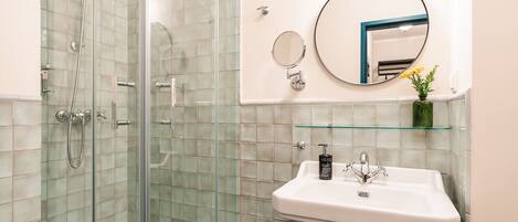 Un baño con cabina de ducha de vidrio, un lavabo blanco con un espejo encima y un pequeño florero verde con flores sobre una estantería.