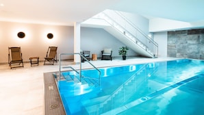A modern indoor swimming pool with a glass barrier, a staircase, and seating areas with chairs and a potted plant.