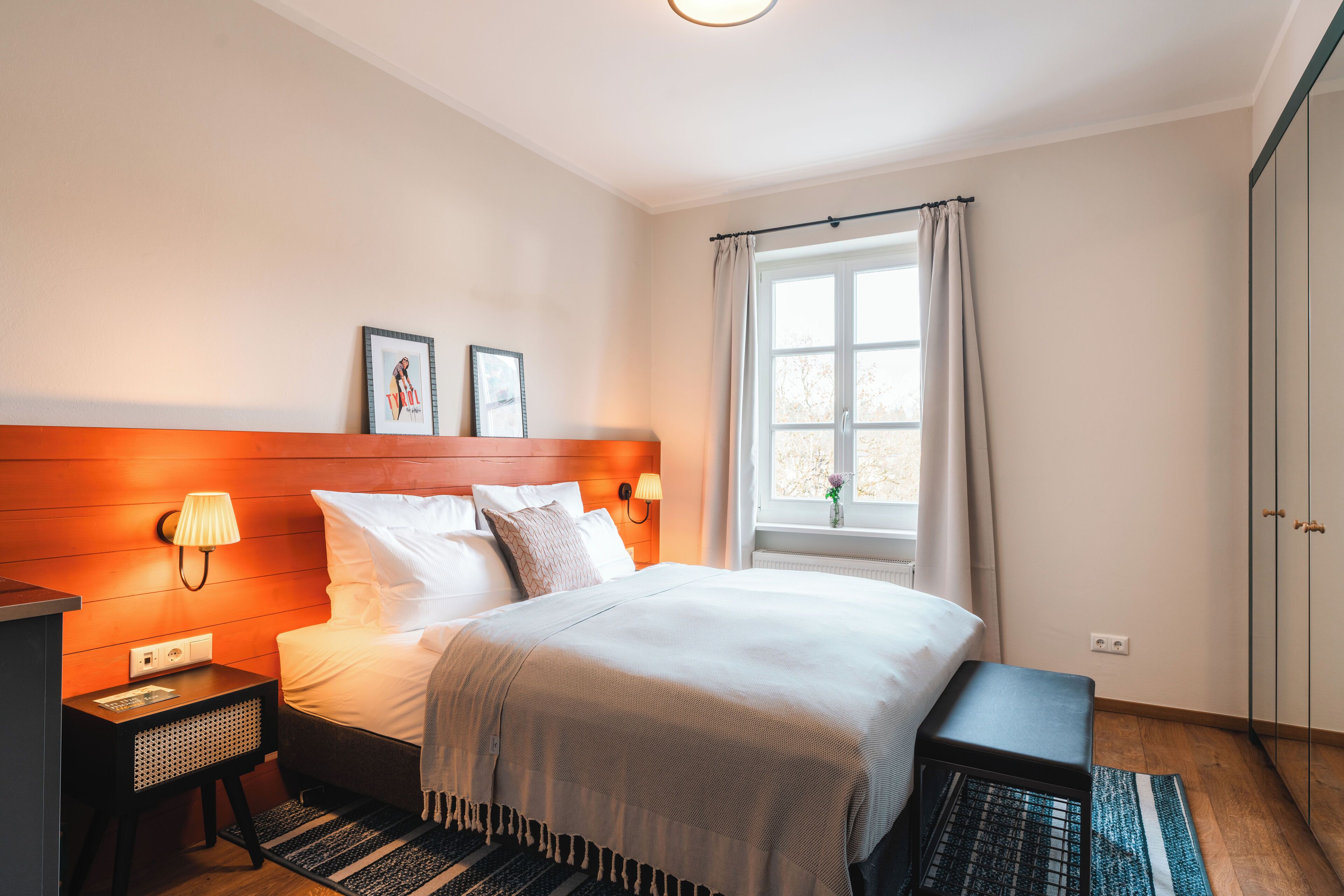 Ein ordentlich eingerichtetes Hotelzimmer mit Bett, Nachttischen, einem Fenster mit Vorhängen und einem Kleiderschrank.