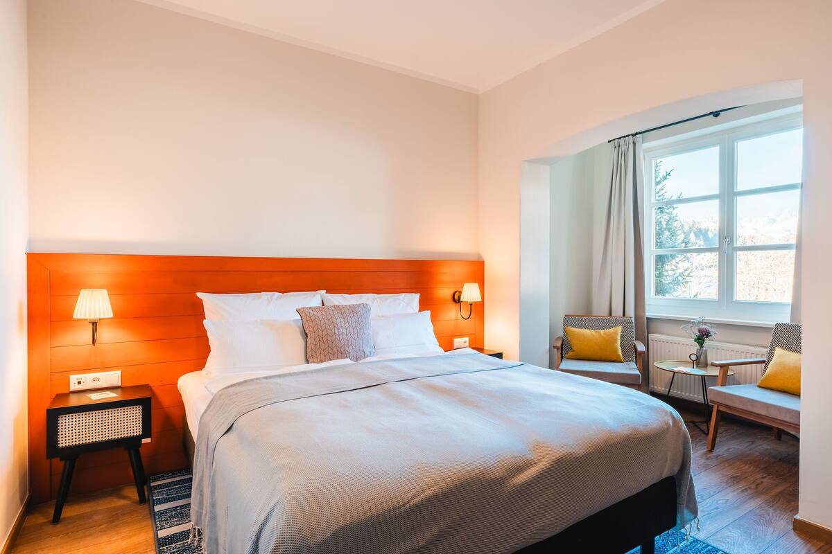 A hotel room with a large bed, two bedside lamps, a small table with a chair, and a window with curtains.