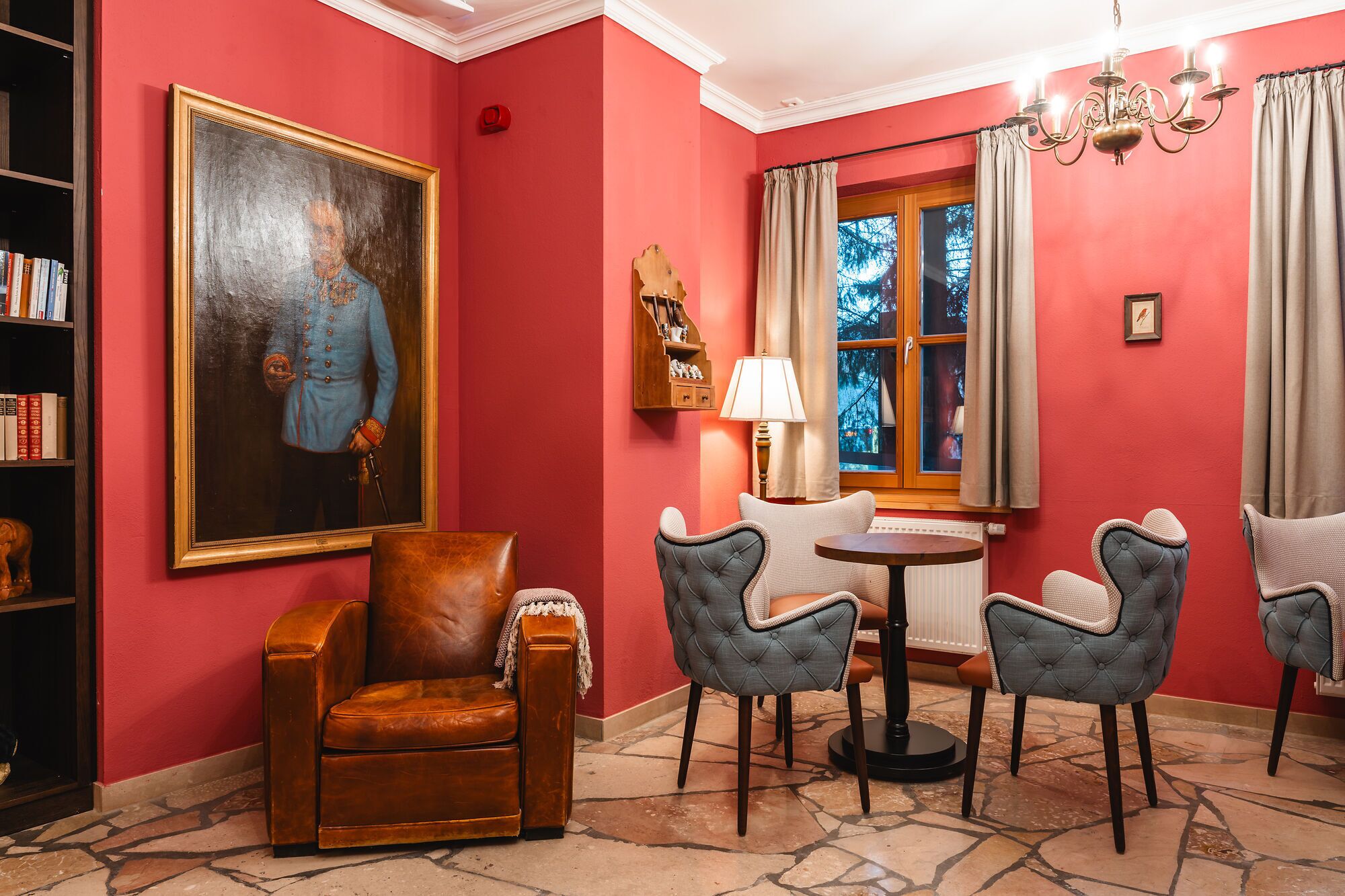 A room with a red wall, a large portrait, a leather armchair, two tufted chairs, a small table, a lamp, and a window with curtains.