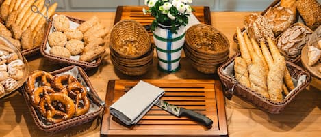 A variety of breads and pastries arranged on a wooden table with a knife and a vase of flowers in the center.