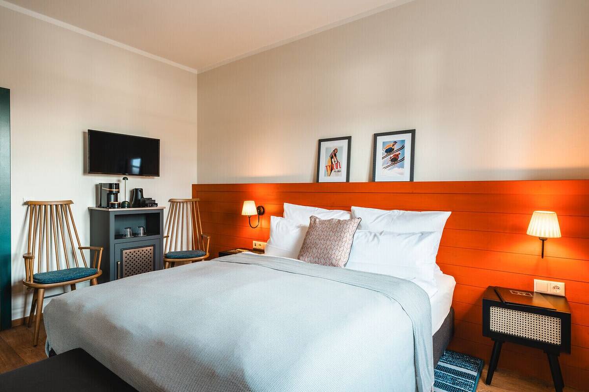 A hotel room with a bed, a TV, a desk with a coffee maker, two chairs, and framed pictures on the wall.