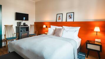 A hotel room with a bed, a TV, a desk with a coffee maker, two chairs, and framed pictures on the wall.