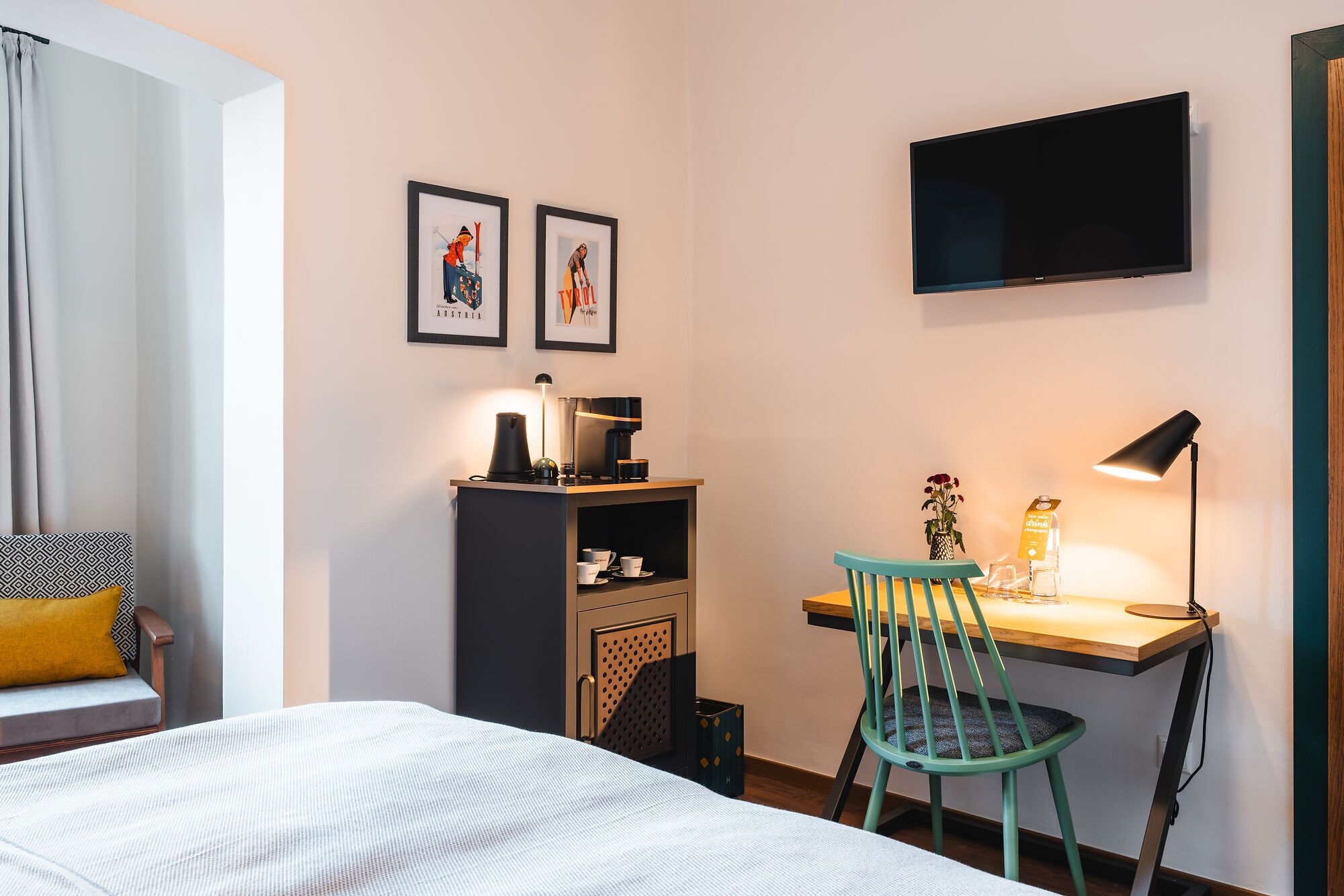 A hotel room with a bed, a desk with a chair, a coffee machine, a TV, and two framed pictures on the wall.