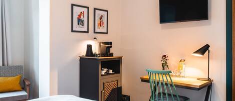 A hotel room with a bed, a desk with a chair, a coffee machine, a TV, and two framed pictures on the wall.