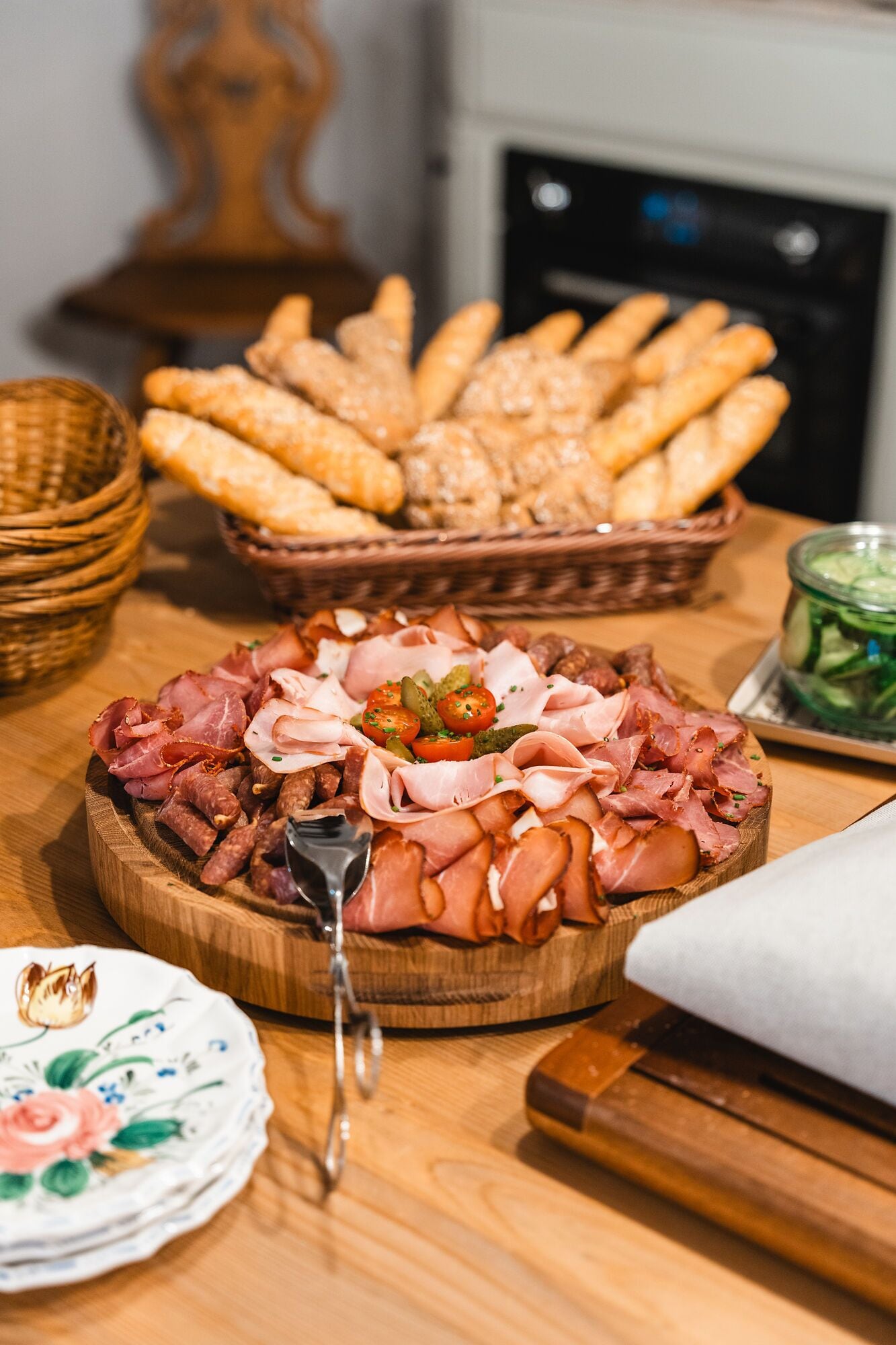 A wooden board with various meats and a basket of breadsticks.