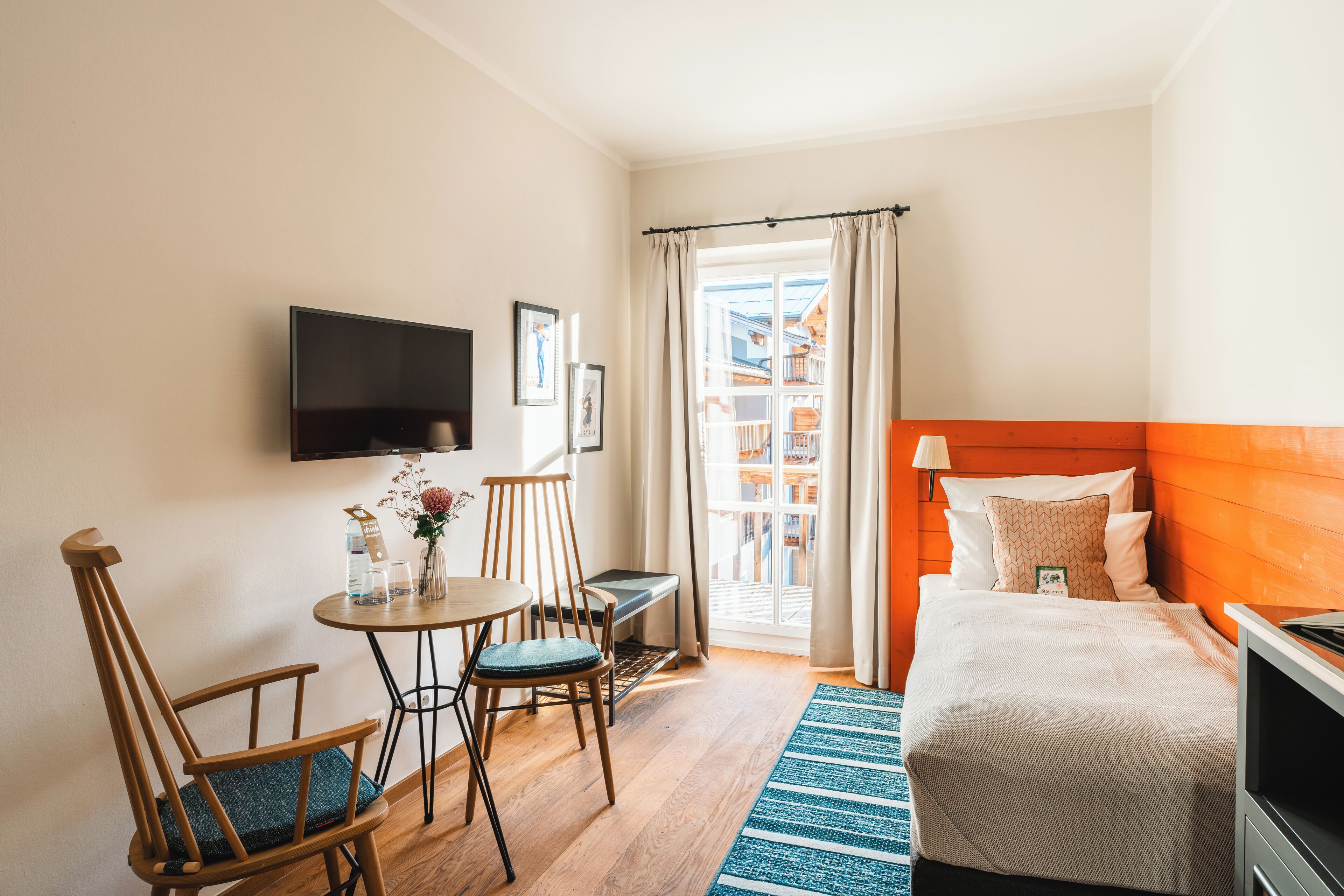 A hotel room with a bed, a TV mounted on the wall, a small table with chairs, and a window with curtains.