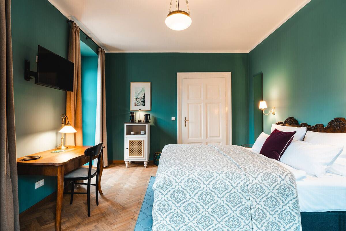 A bedroom with teal walls, a bed with patterned bedding, a desk with a lamp, a television, and a door.