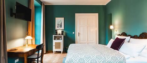A bedroom with teal walls, a bed with patterned bedding, a desk with a lamp, a television, and a door.