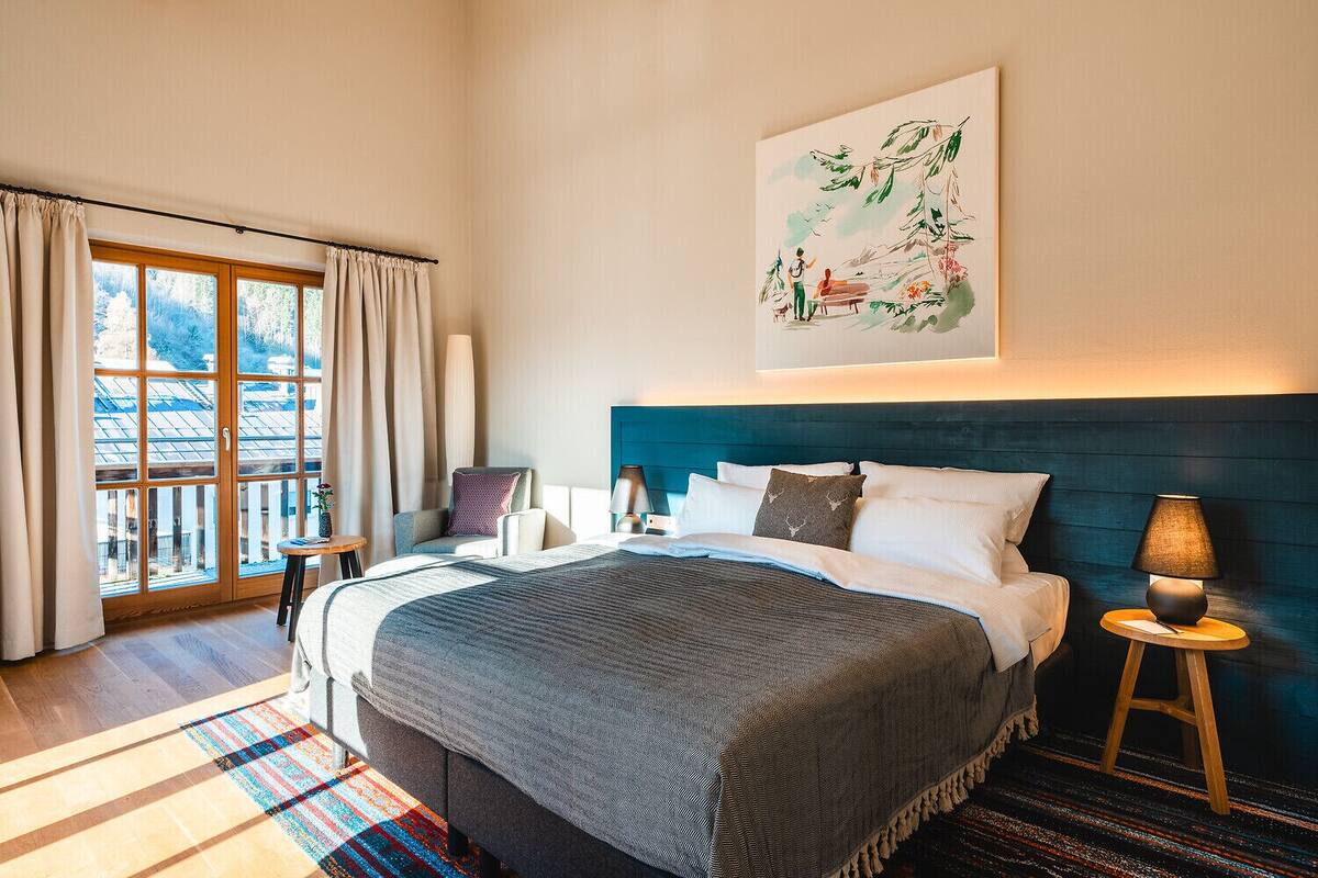 A modern hotel room with a large bed, a view of the outdoors through a sliding glass door, a painting on the wall, and a small table with a lamp.
