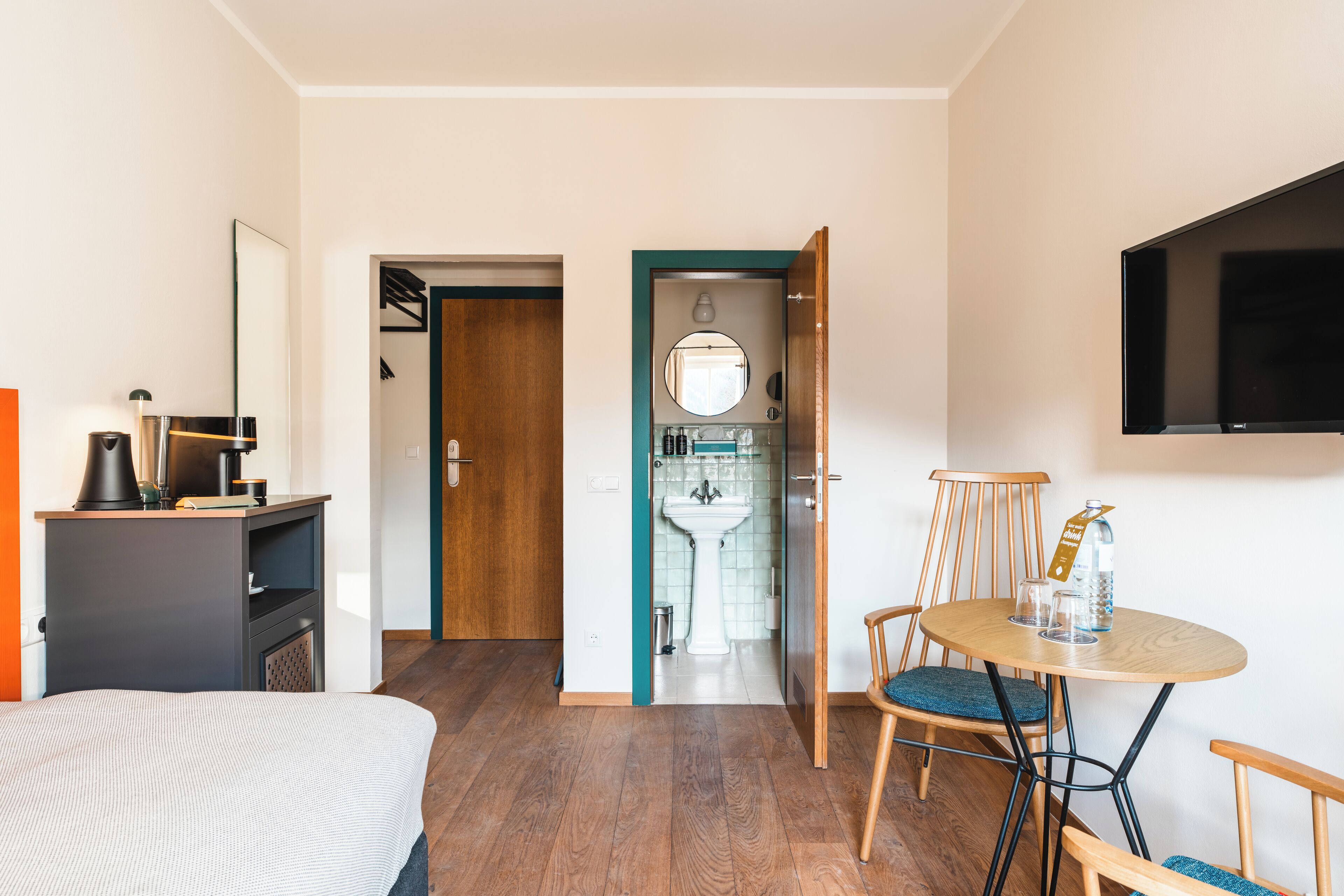Ein Hotelzimmer mit einem Bett, einem Schreibtisch mit Lampe, einem runden Tisch mit Stühlen, einem an der Wand befestigten Fernseher und einem durchgegangenen Tür erkennbaren Badezimmer.