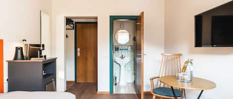 A hotel room with a bed, a desk with a lamp, a round table with chairs, a TV mounted on the wall, and a bathroom visible through an open door.