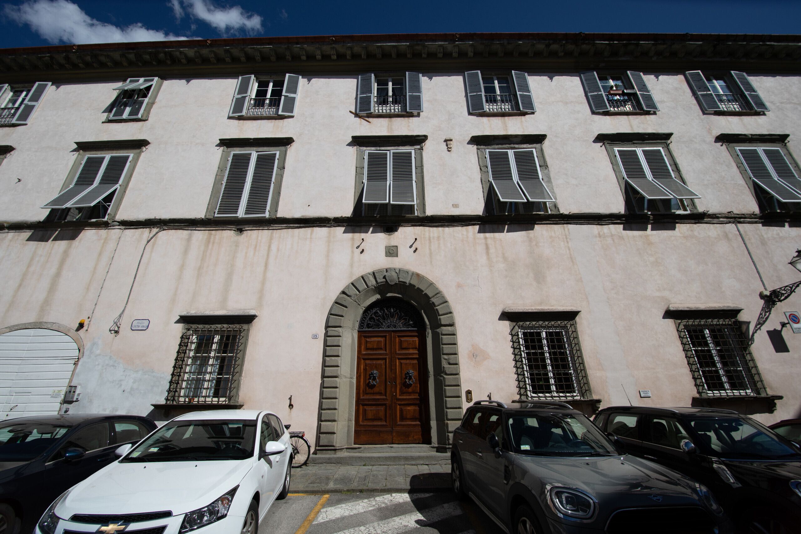 A building with a central arched entrance, multiple windows with shutters, and parked cars in front.