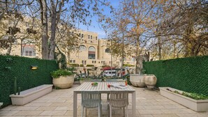 Une terrasse extérieure avec une table et des chaises, entourée de verdure et avec un bâtiment en arrière-plan.