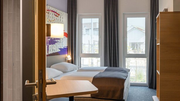 Habitación de hotel con cama, escritorio y silla. Dos ventanas con cortinas y vistas a edificios.
