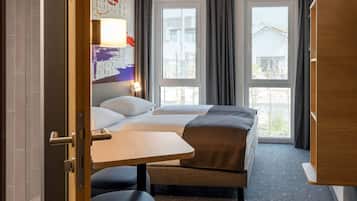 Habitación de hotel con cama, escritorio y silla. Dos ventanas con cortinas y vistas a edificios.