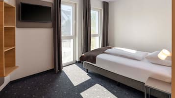 A hotel room with a bed, a TV mounted on the wall, a window with curtains, and a small table with a lamp.