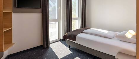 A hotel room with a bed, a TV mounted on the wall, a window with curtains, and a small table with a lamp.