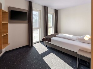 Una habitación de hotel con una cama, un televisor montado en la pared, una ventana con cortinas y una pequeña mesa con una lámpara.