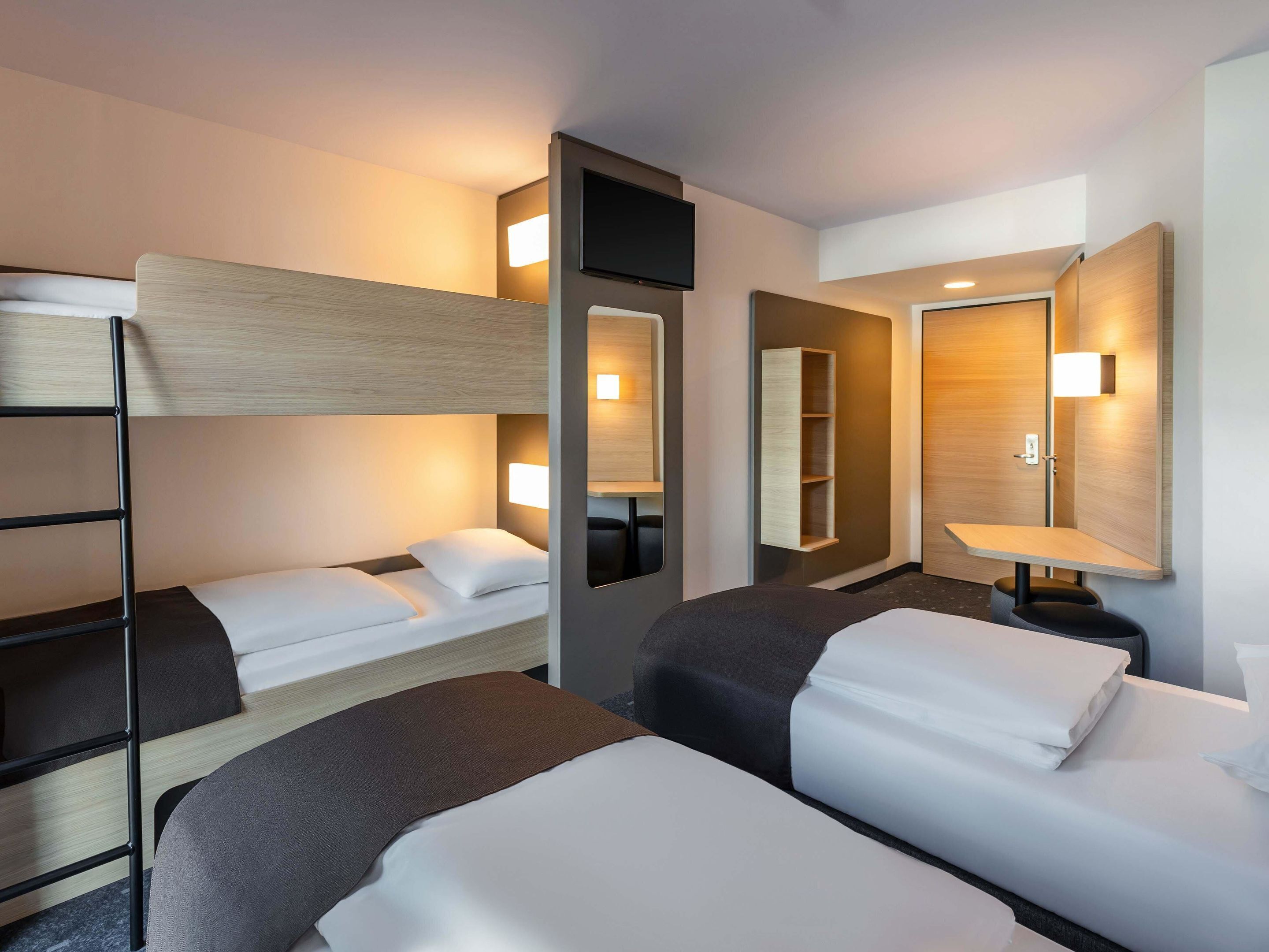 Una habitación de hotel con literas, una mesita de noche con lámpara, un espejo y un estante en la pared.