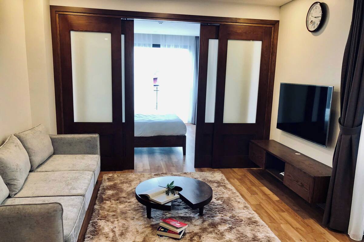Una habitación de hotel moderna con un sofá, una mesa de centro con libros, un televisor y un ventanal con cortinas.