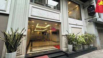 Entrada de un edificio con fachada de mármol, un felpudo que dice 'Welcome' y plantas en macetas.