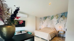A modern bedroom with a bed, a desk with a chair, a TV, and a decorative wall with a tropical mural.
